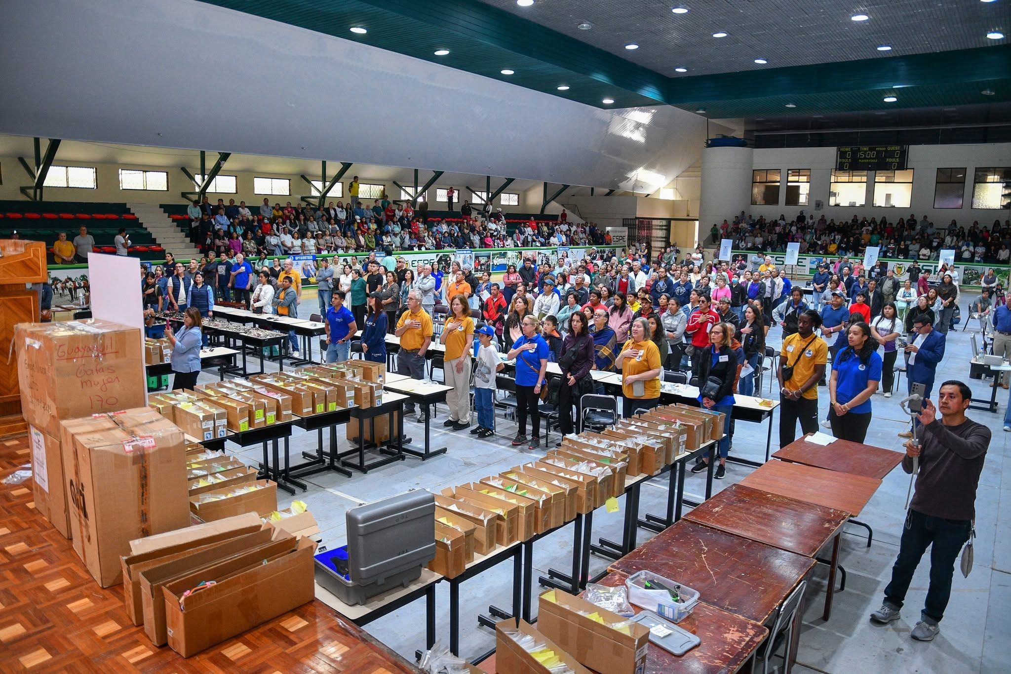 Coliseo lleno en la apertura de la campaña: decenas de mesas con cajas de lentes reciclados clasificados por medida, el público de pie y los voluntarios del club atendiendo las filas.
